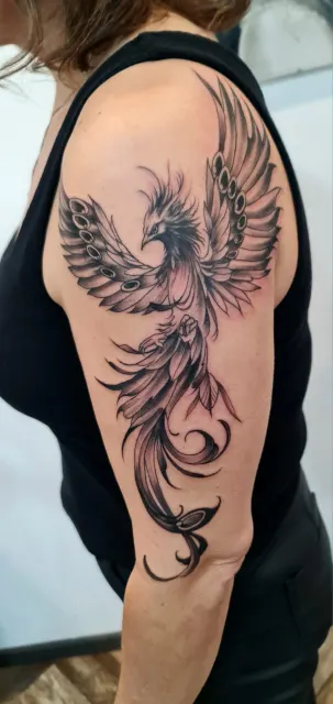 Rozbudowany czarno-szary tatuaż feniksa wykonany na ramieniu. Projekt wykorzystuje kształt ręki i łączy wyraziste skrzydła z dekoracyjnymi detalami. Realizacja One More Tattoo w Skarżysku-Kamiennej.